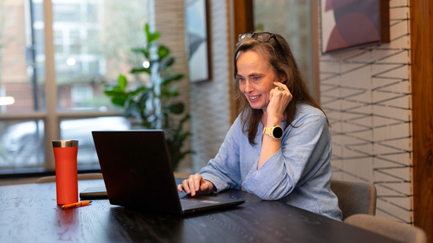 Person seated at a table, using a laptop while holding a phone, with a travel mug nearby in a modern indoor space.