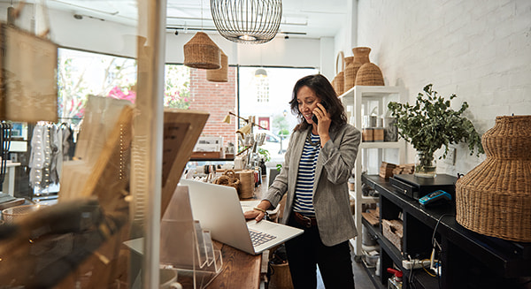A woman in a small boutique shop talks on the phone while working on a laptop at a wooden counter surrounded by woven baskets and home decor items