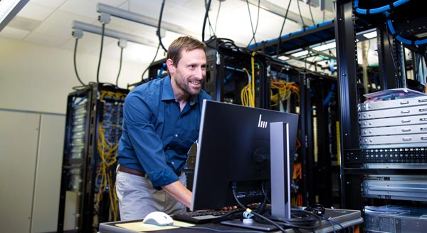 Person working at a desktop computer in a server room, surrounded by racks of networking equipment and cables.