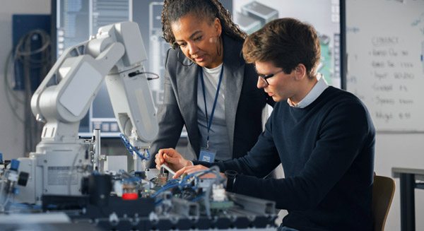 Two people working together on an electronics project beside a robotic arm in a lab.