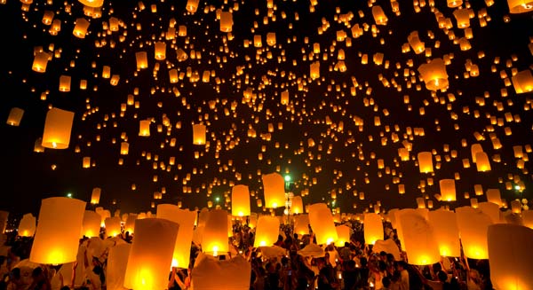 Hundreds of glowing lanterns rise into the night sky during a large cultural festival, with a crowd of participants releasing lanterns together.