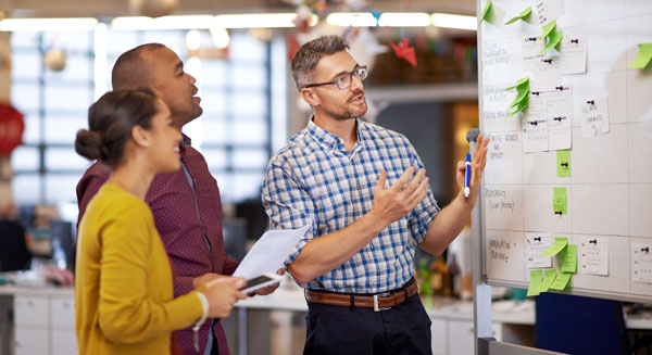 Three people collaborate in an open office, reviewing notes and sticky‑notes on a large whiteboard during a planning or research discussion.
