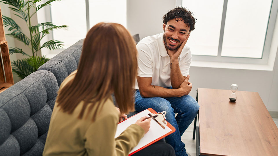 Two people sitting side-by-side in a counseling session. One person is taking notes while another smiles.