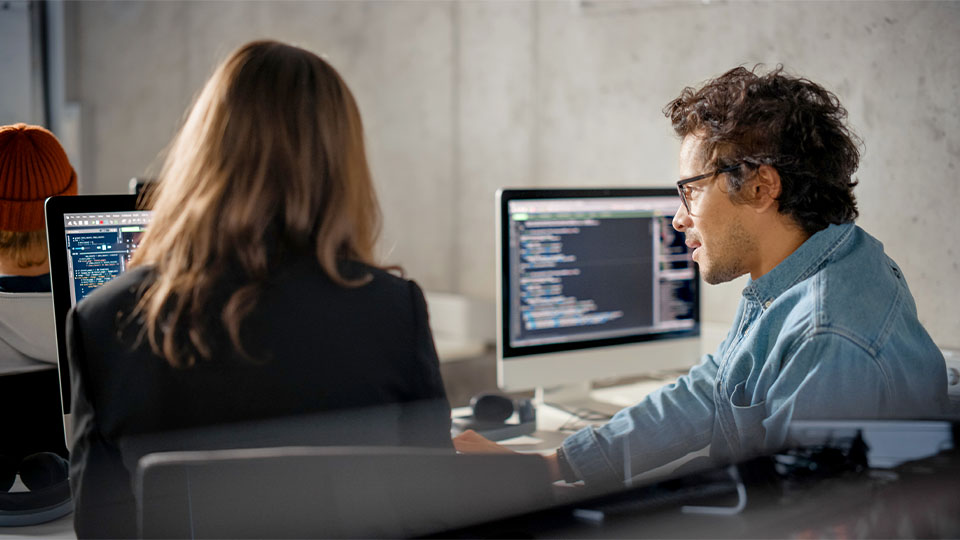 Two computer monitors display code, and a man and woman sit in front of one while discussing what's on screen
