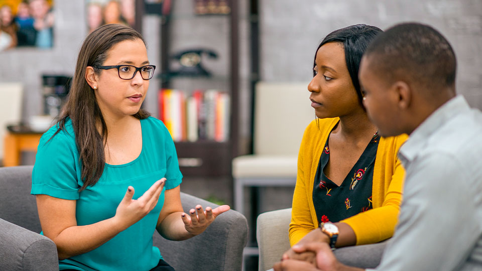 In a private office, a woman wearing glasses sits in a plush chair while counseling two people who sit across from her