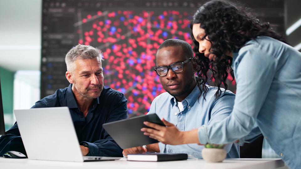 Three people gather around a desk in a workspace environment. One person with gray hair and a casual, long-sleeved shirt is looking at a laptop screen. In the center, a person with glasses and short hair is concentrating on a tablet. On the right, a person with long curly hair is leaning over, pointing at the tablet, and appears to be explaining something.