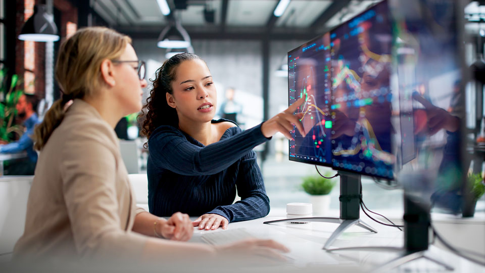 Two people sit at a desk in a modern office environment, focusing on multiple monitors displaying complex graphs and data visuals. One person points at the screen, seemingly explaining the data to the other person.