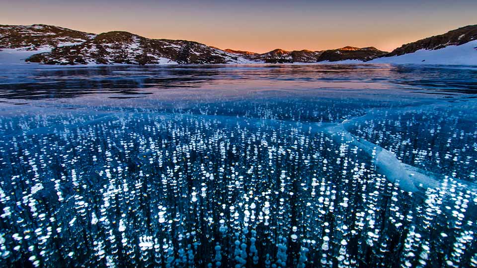 Sunset over a frozen lake with bubbles trapped in the ice and snow-covered hills in the background.