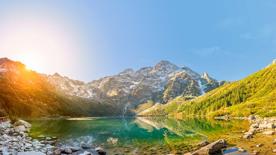 In a panoramic view of Morskie Oko, a lake in the Polish Tatra Mountains, the sun crests over a rocky peak behind the lake