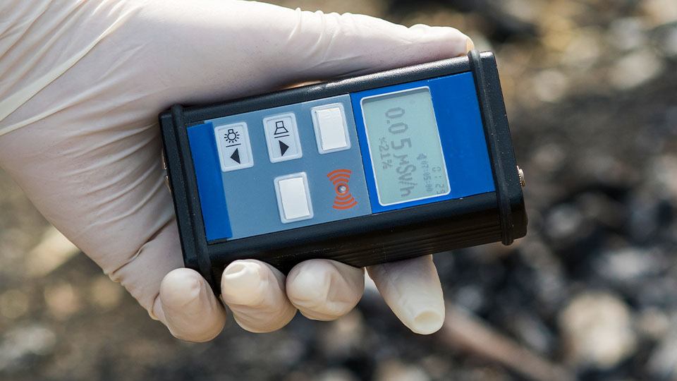 Close-up of a portable radiation monitoring device held in a gloved hand, with a digital screen displaying measurement values.