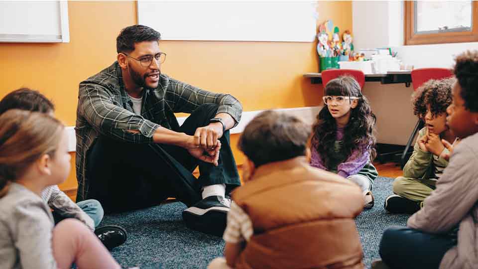 A male teacher sits on the floor with young students in a circle, engaging them in discussion in a colorful classroom setting