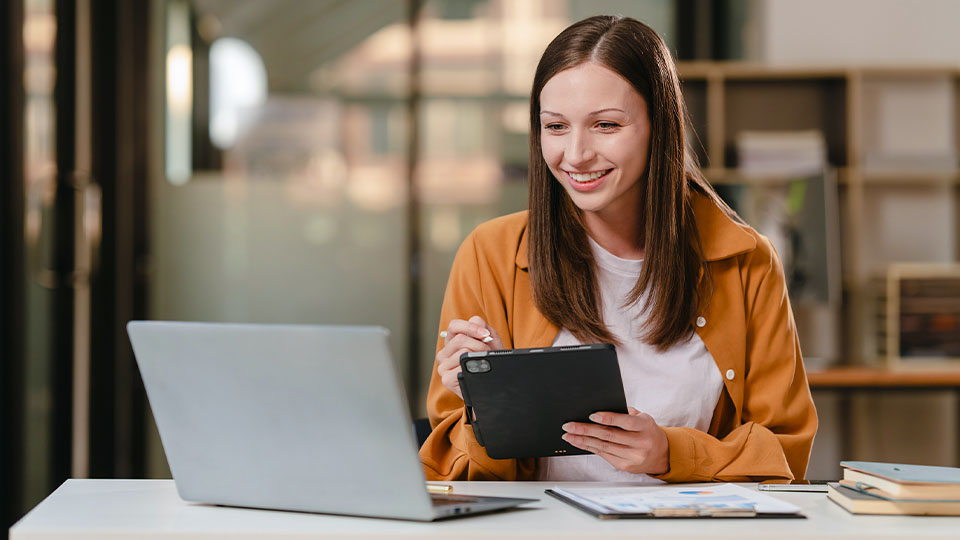 Smiling woman using a tablet and laptop in an office, analyzing data to explore AI applications in psychology.