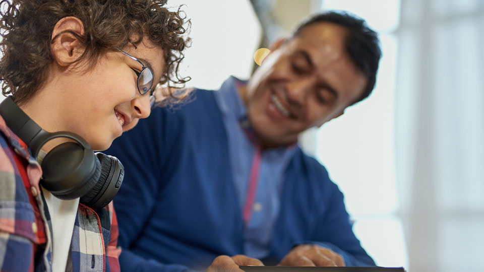 Smiling adult supports a curly-haired young person wearing glasses and headphones while using a digital device in a bright setting.