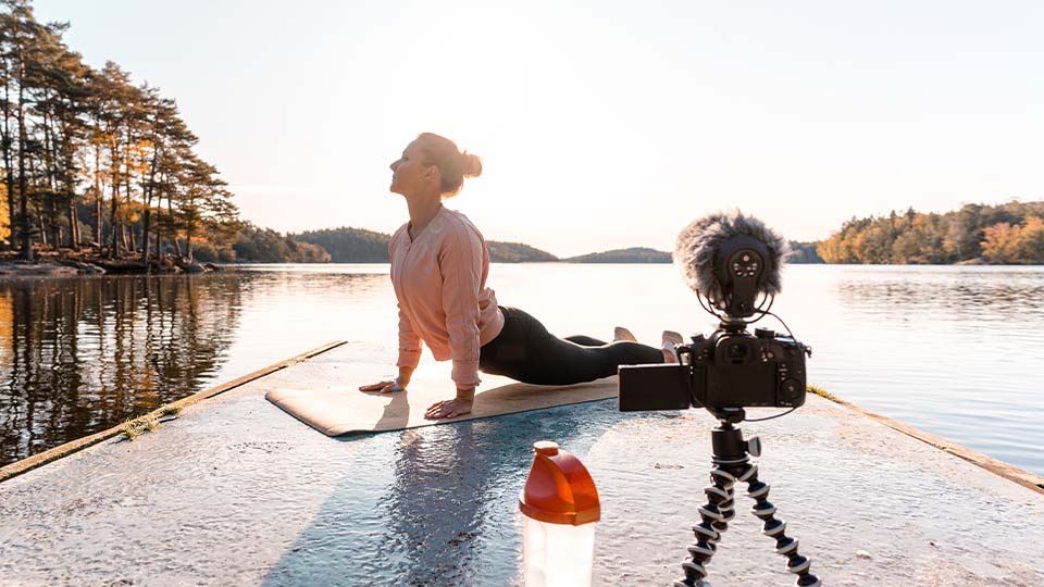Person performing an upward-facing yoga pose on a mat at the edge of a dock overlooking a calm lake, with a camera on a tripod and a pink water bottle in the foreground, surrounded by trees under a clear sky.