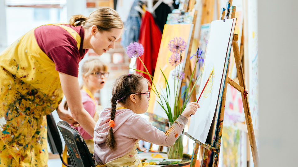 Art instructor assisting a child painting on an easel in a bright studio, with colorful flowers in a vase and art supplies scattered around.
