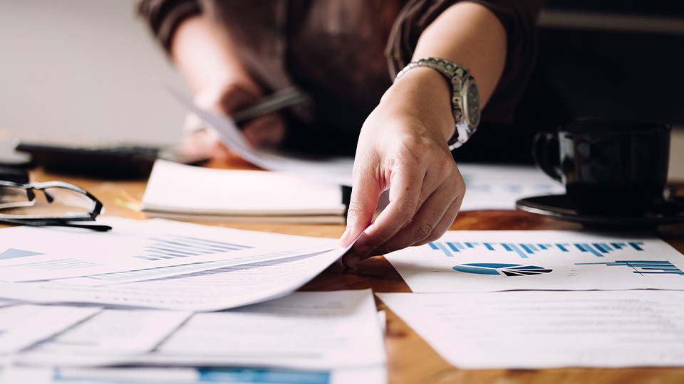 Close-up of a person’s hand sorting through printed charts and graphs on a wooden desk, with a coffee cup, eyeglasses, and documents scattered around.