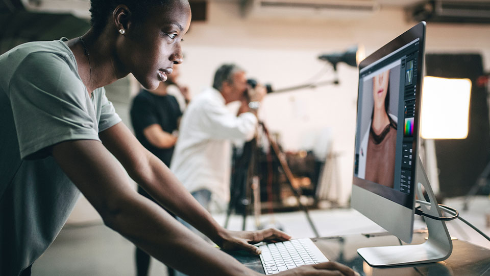 Person working on photo editing software at a desktop computer in a studio, with a camera crew and lighting equipment visible in the background.