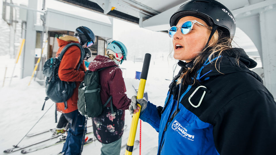 Group of people in winter gear standing under a ski lift structure, holding ski poles and wearing helmets, with snow-covered ground and equipment visible in the background.