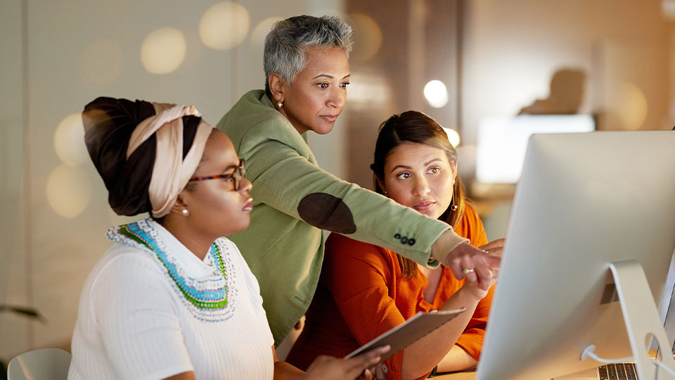 Three people collaborating in an office setting, with one pointing at a large desktop monitor while another holds a tablet, and the third seated nearby.