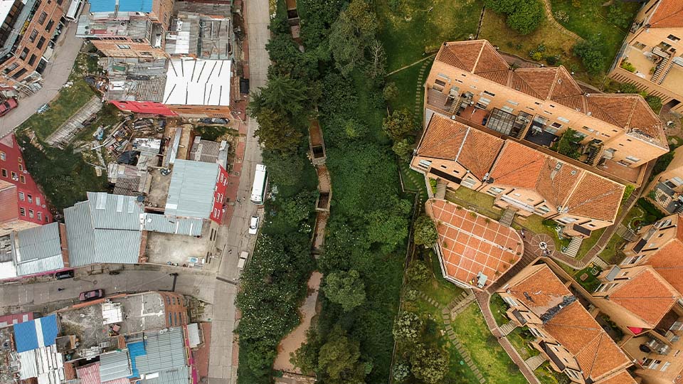 Aerial view showing a stark contrast between two neighborhoods: densely packed homes with metal roofs on the left and well-maintained townhouses with red-tiled roofs on the right, separated by a strip of green trees.
