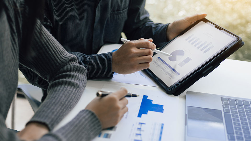 Two people reviewing financial charts and graphs on a tablet, using stylus pens, with printed bar charts and a laptop on the table.