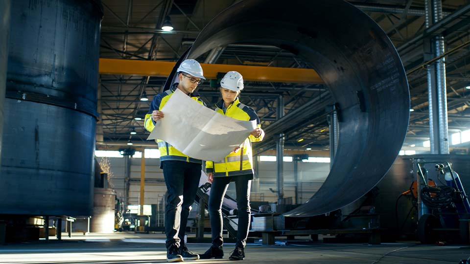 Two people wearing safety helmets and high-visibility jackets examining a large blueprint inside an industrial facility, with massive cylindrical metal structures and overhead cranes in the background.