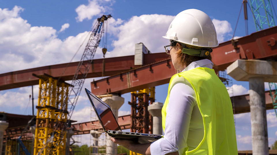 Construction site with a person wearing a hard hat and high-visibility vest holding an open laptop, observing steel beams and support columns being assembled under a partly cloudy sky.
