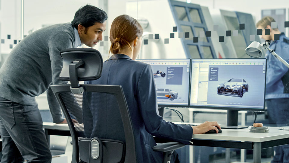 Modern office environment with multiple people working at computer stations; two monitors display 3D car design models, while another person stands in the background near industrial equipment.