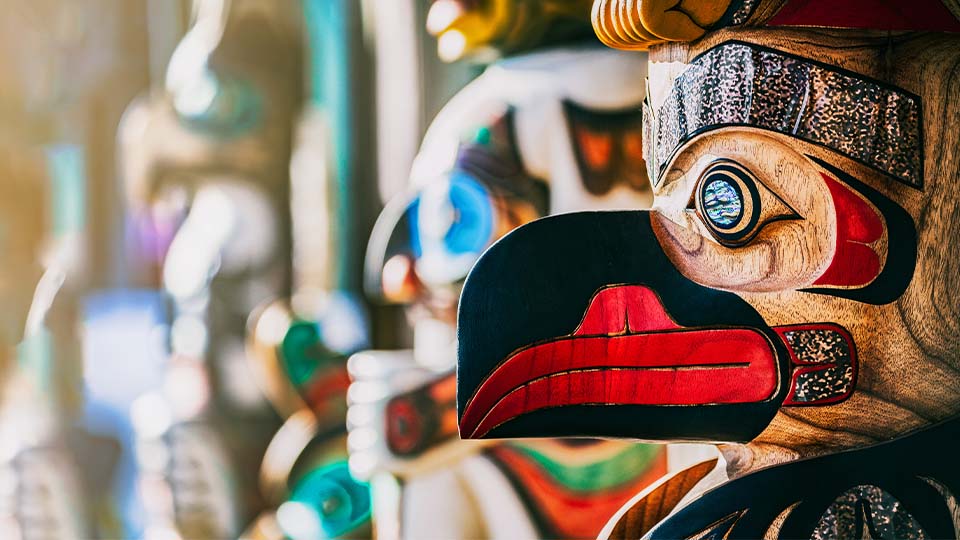 Close-up of a traditional Indigenous totem pole featuring a brightly painted bird figure with bold red, black, and turquoise details, set against a softly blurred background of other carved figures.