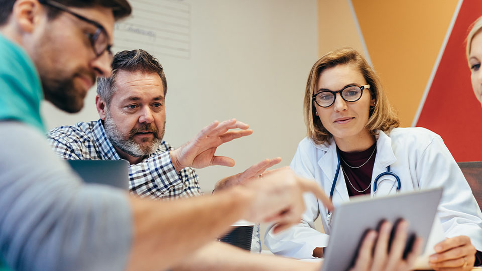 Group of people in a meeting, with one person in a white coat and stethoscope holding a tablet while others discuss and gesture toward the screen, seated around a table in a brightly lit room.