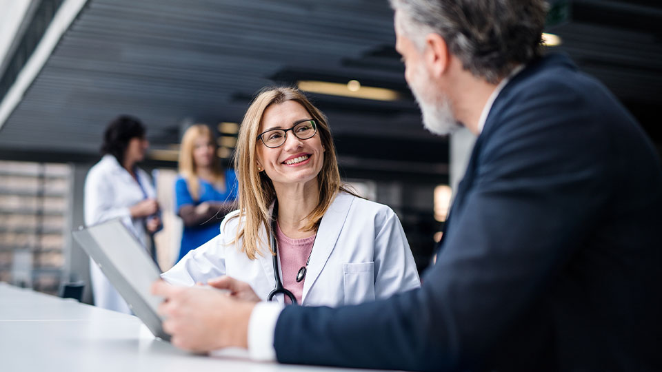 Healthcare professional in a white coat holding a tablet and speaking with a person in business attire across a table, with two additional people in similar white coats conversing in the background inside a modern, well-lit building.