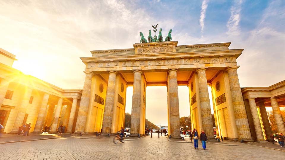 The Brandenburg Gate in Berlin, Germany, shown in warm sunlight with a clear blue sky and scattered clouds.