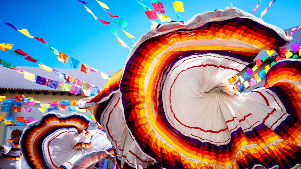 A vibrant outdoor scene featuring dancers in traditional Mexican attire, with large flowing skirts in white accented by bold stripes of orange, purple, and yellow.