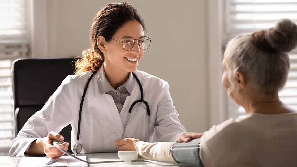 A healthcare professional wearing a white coat and stethoscope sits at a desk speaking with an older person who has gray hair tied in a bun.
