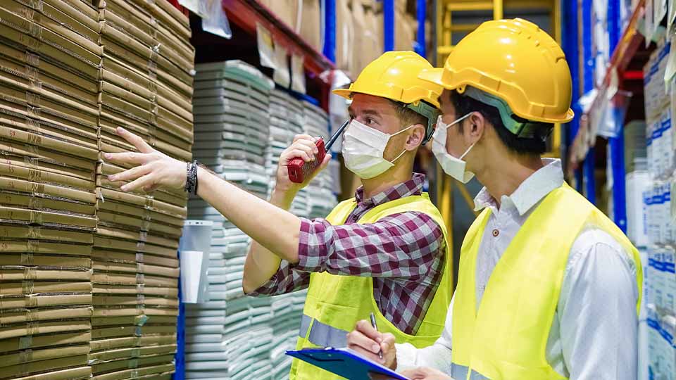 Two workers wearing yellow safety vests and hard hats stand in a warehouse aisle. One worker is pointing toward a tall stack of packaged goods while holding a walkie-talkie, and the other is holding a blue clipboard and pen.