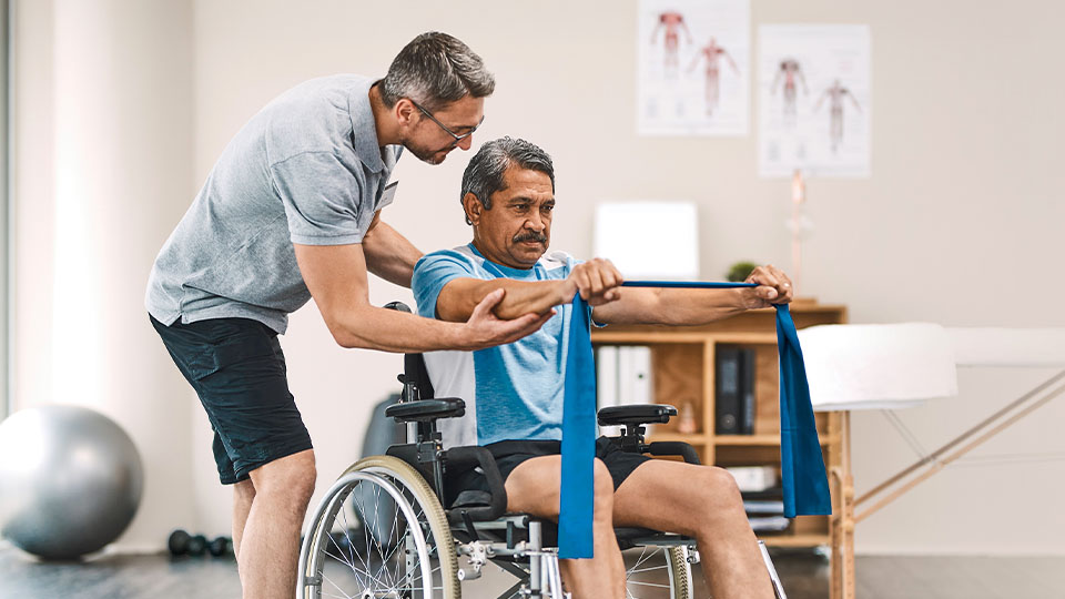 A person in a wheelchair performs an arm exercise using a blue resistance band while another person assists by supporting the arm.