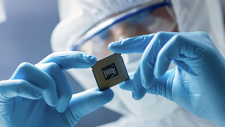A person wearing blue gloves holds a small computer processor chip with visible gold contacts and a central grid pattern, in a cleanroom environment with protective gear and soft lighting.