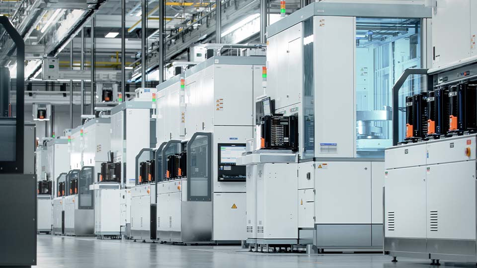 A row of advanced industrial machines inside a clean, modern manufacturing facility. The equipment is white with glass panels and orange accents, arranged in a well-lit space with overhead piping and cables visible.