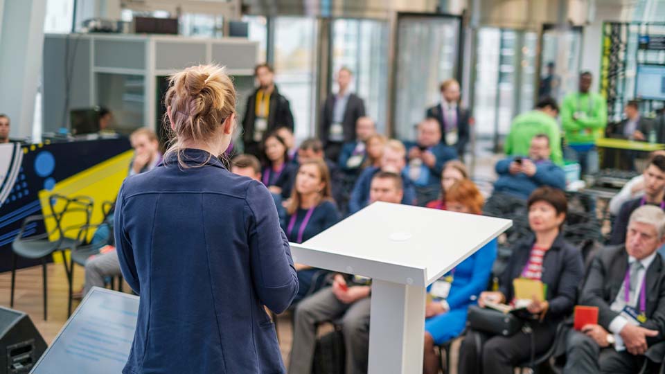 A person stands at a white podium in a modern conference space, addressing an audience seated in rows.
