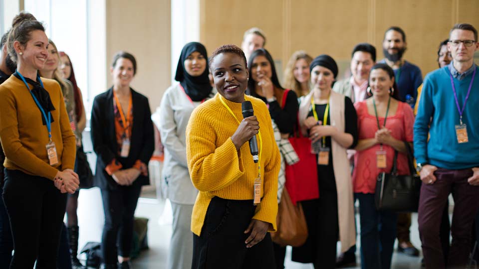 A person wearing a bright yellow sweater is standing and holding a microphone while speaking to a group of people in a well-lit indoor setting.