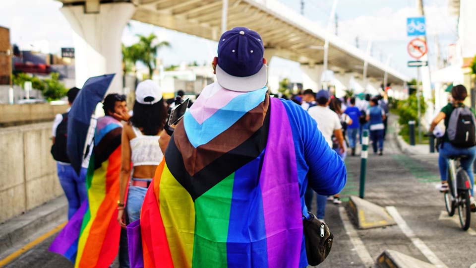 A group of people walking along a city street during a public event or march. The central figure has a large rainbow-colored Pride flag draped over their shoulders.