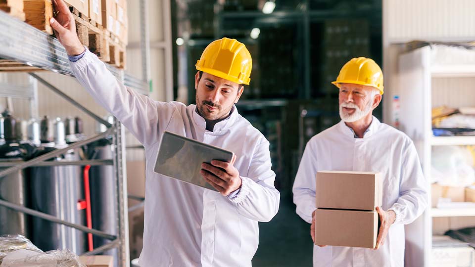 Two workers wearing yellow safety helmets and white shirts are in a warehouse setting. One person is holding a tablet and reaching toward items stored on a high shelf, while the other carries two cardboard boxes.