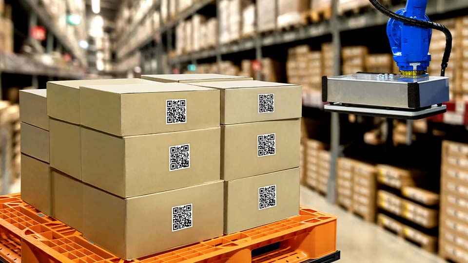A stack of cardboard boxes with QR codes is placed on an orange pallet in a warehouse aisle. In the foreground, a robotic arm with a blue gripper is positioned near the boxes.