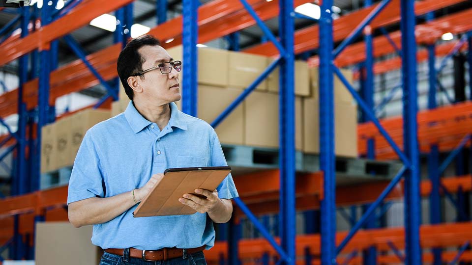 A person in a light blue collared shirt is holding a tablet while standing in a warehouse. Behind them are tall blue and orange metal racks stacked with cardboard boxes.