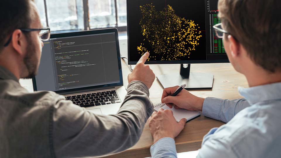 Two people are seated at a desk in front of computer screens, collaborating on a data analysis project.