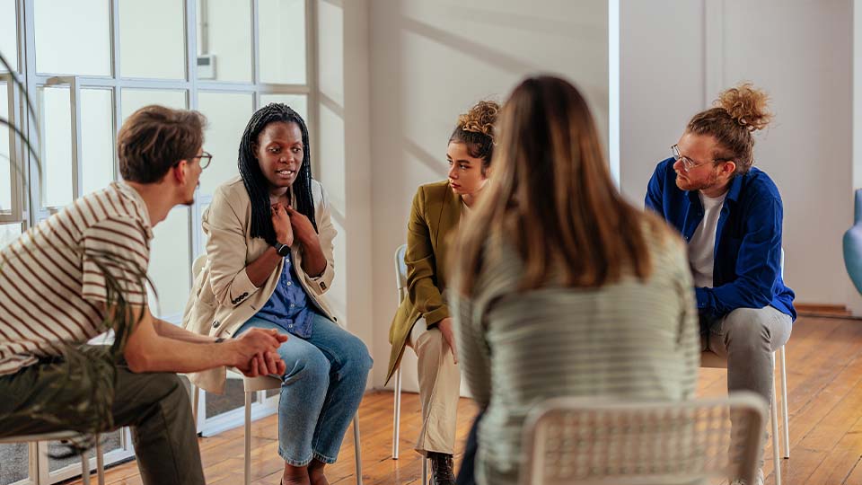 Woman speaks to a small group seated in a circle during a thoughtful discussion in a bright, sunlit room