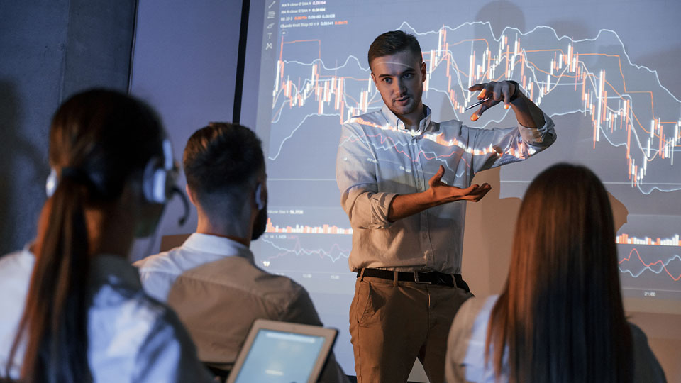 A man presents financial data to a group, gesturing in front of a screen displaying complex charts