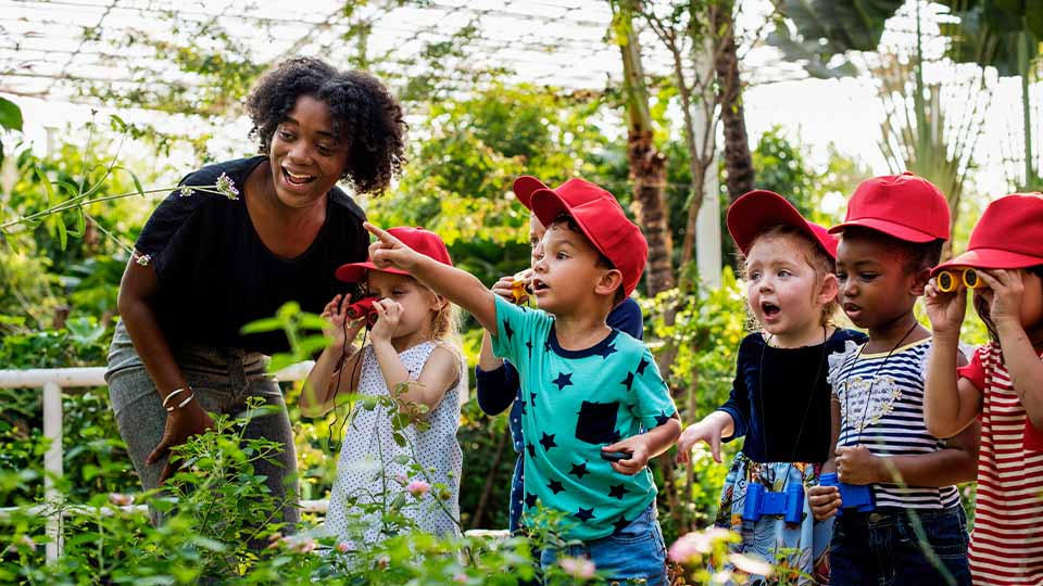 Elementary education teacher guiding young children on an outdoor nature activity, with kids wearing red hats and using binoculars