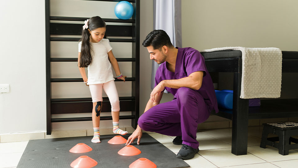 A physical therapist in purple scrubs guides a young girl through a cone exercise