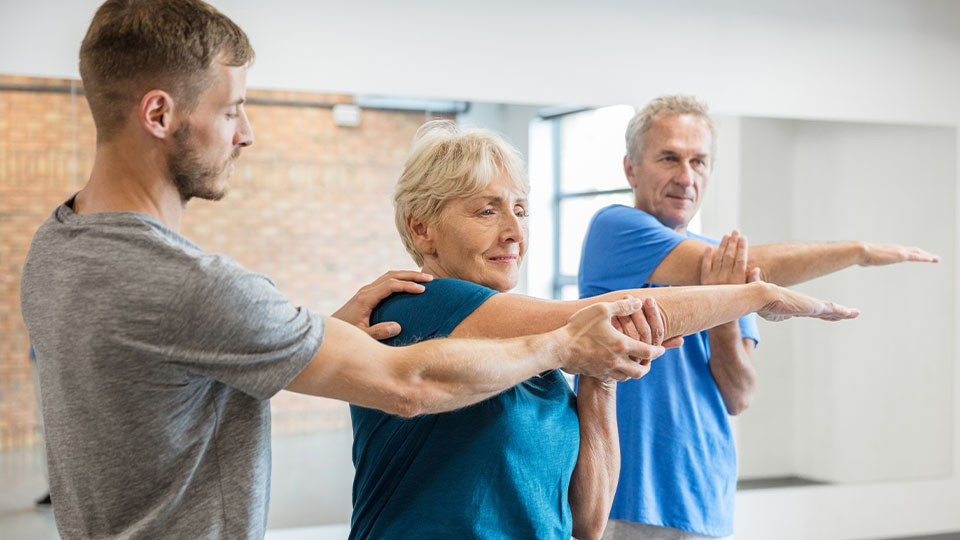 Older couple stretching their upper torsos with support from a younger male physical therapist